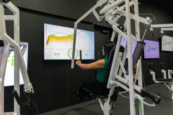 A person using gym equipment with a large screen in front of them, displaying fitness data using JouleFit technology.