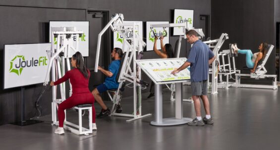 A fitness studio features several strength‑training machines arranged in a row, each branded with the JouleFit logo displayed on nearby monitors. Multiple individuals are using the equipment, including seated resistance machines and a leg‑press setup. One person stands at a large interactive control panel or touchscreen kiosk in front of the machines.